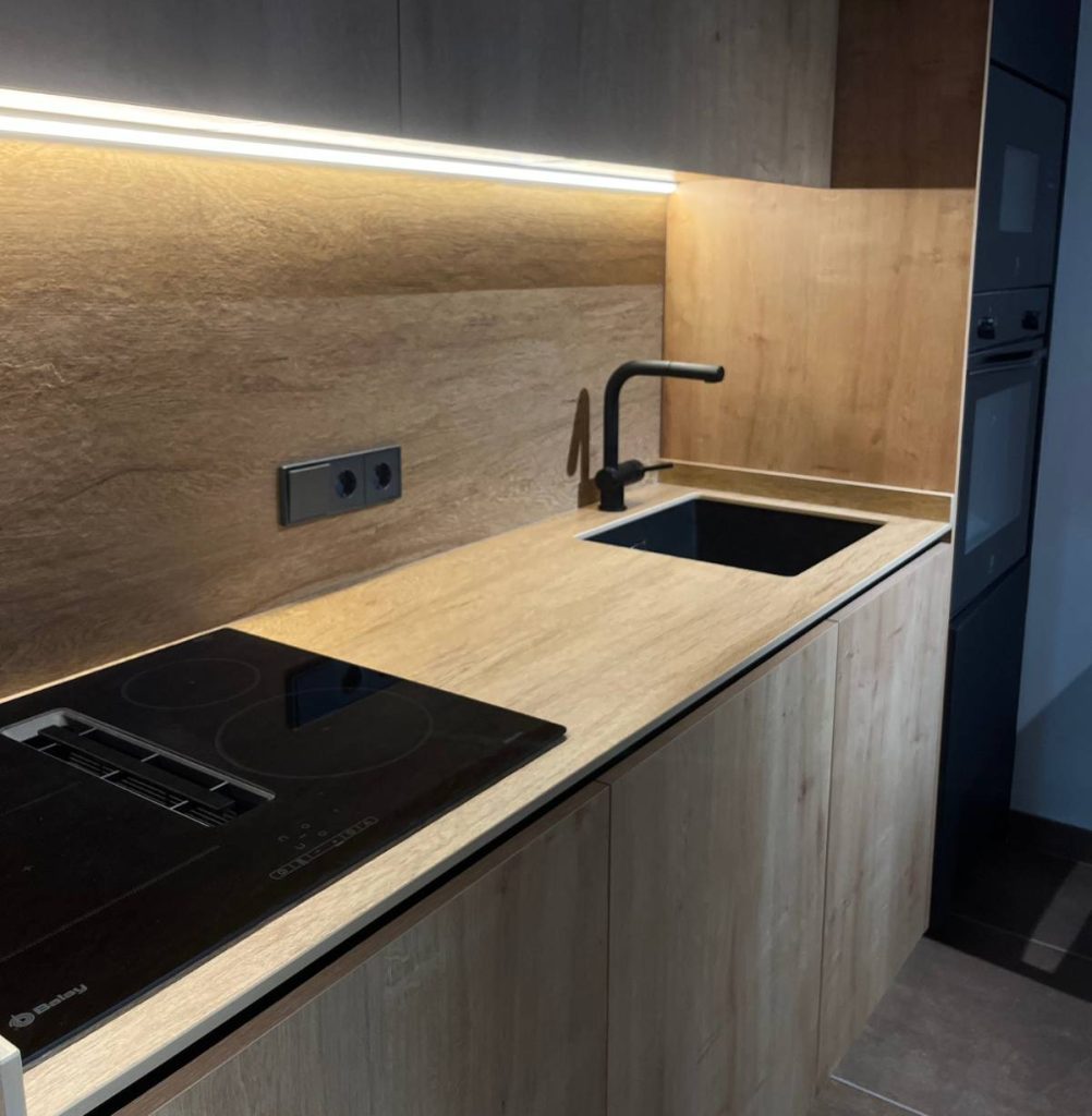 Zona de cuina d’oficina amb mobles de fusta, placa d’inducció, pica integrada i llum LED lineal